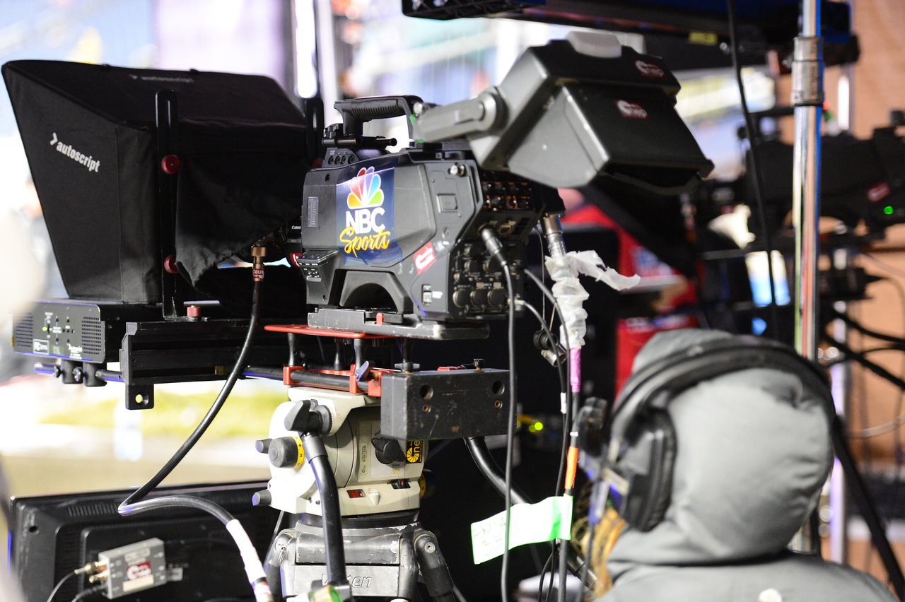 A camera operator wearing headphones films the Patriots game using an NBC Sports broadcast camera with a teleprompter.