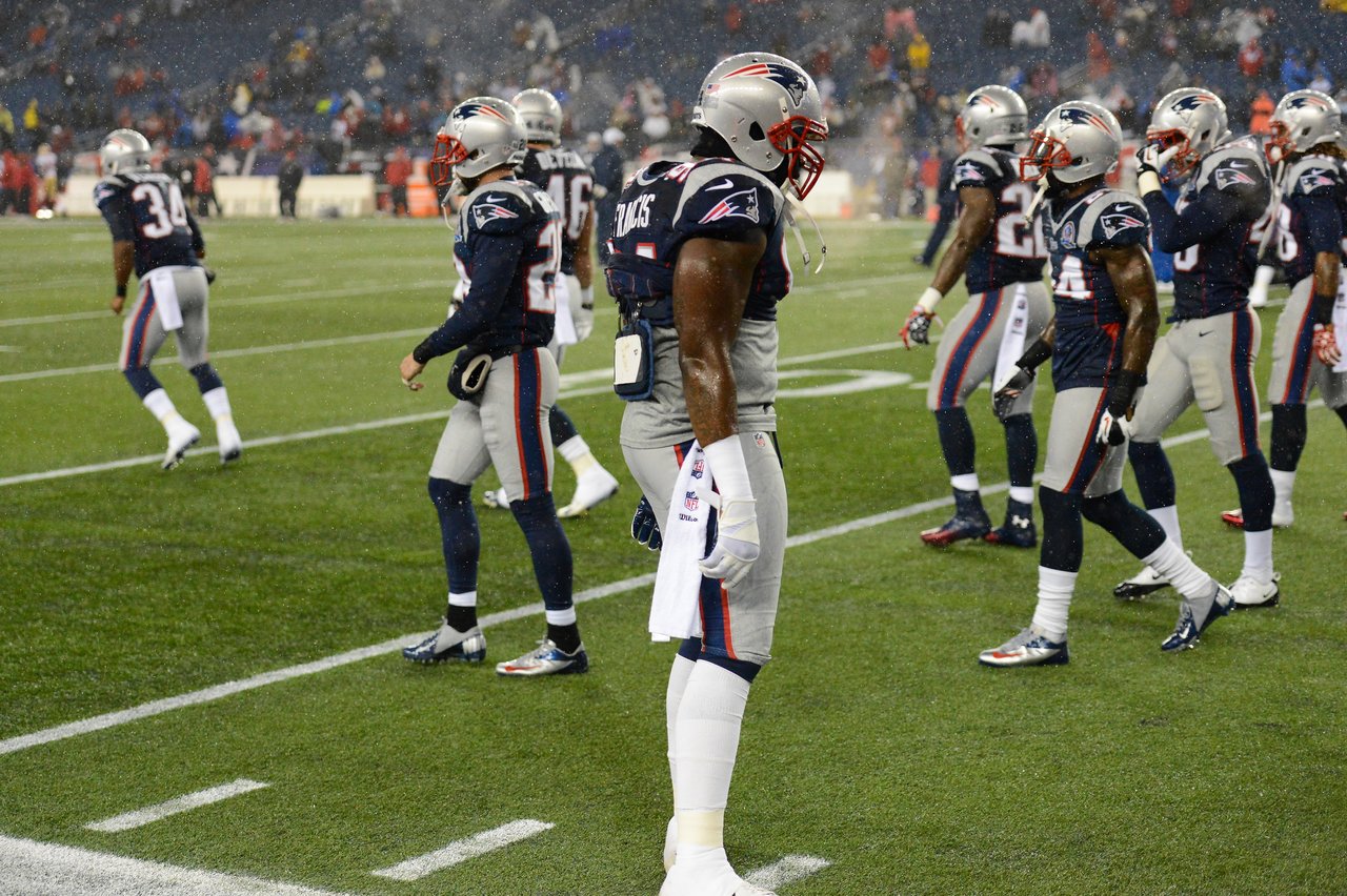 New England Patriots players stand on the field in uniform during a game in the rain.