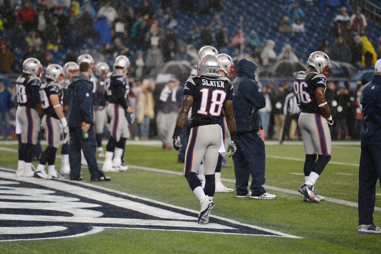 New England Patriots players stand on the sideline during a rainy game, wearing helmets and uniforms.