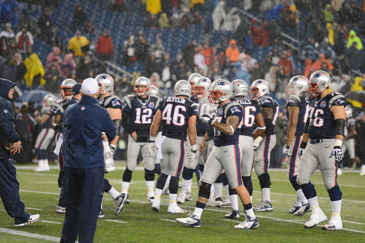 New England Patriots players huddle on the field during a rainy game, wearing helmets and uniforms.