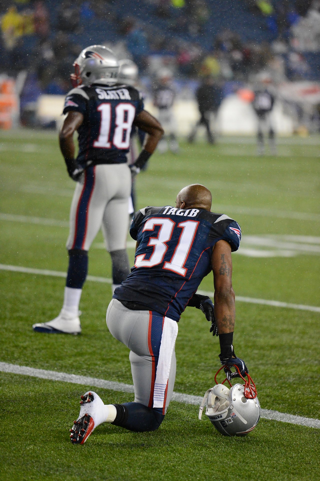 A Patriots player kneels on the field holding his helmet, while another stands nearby during the game.