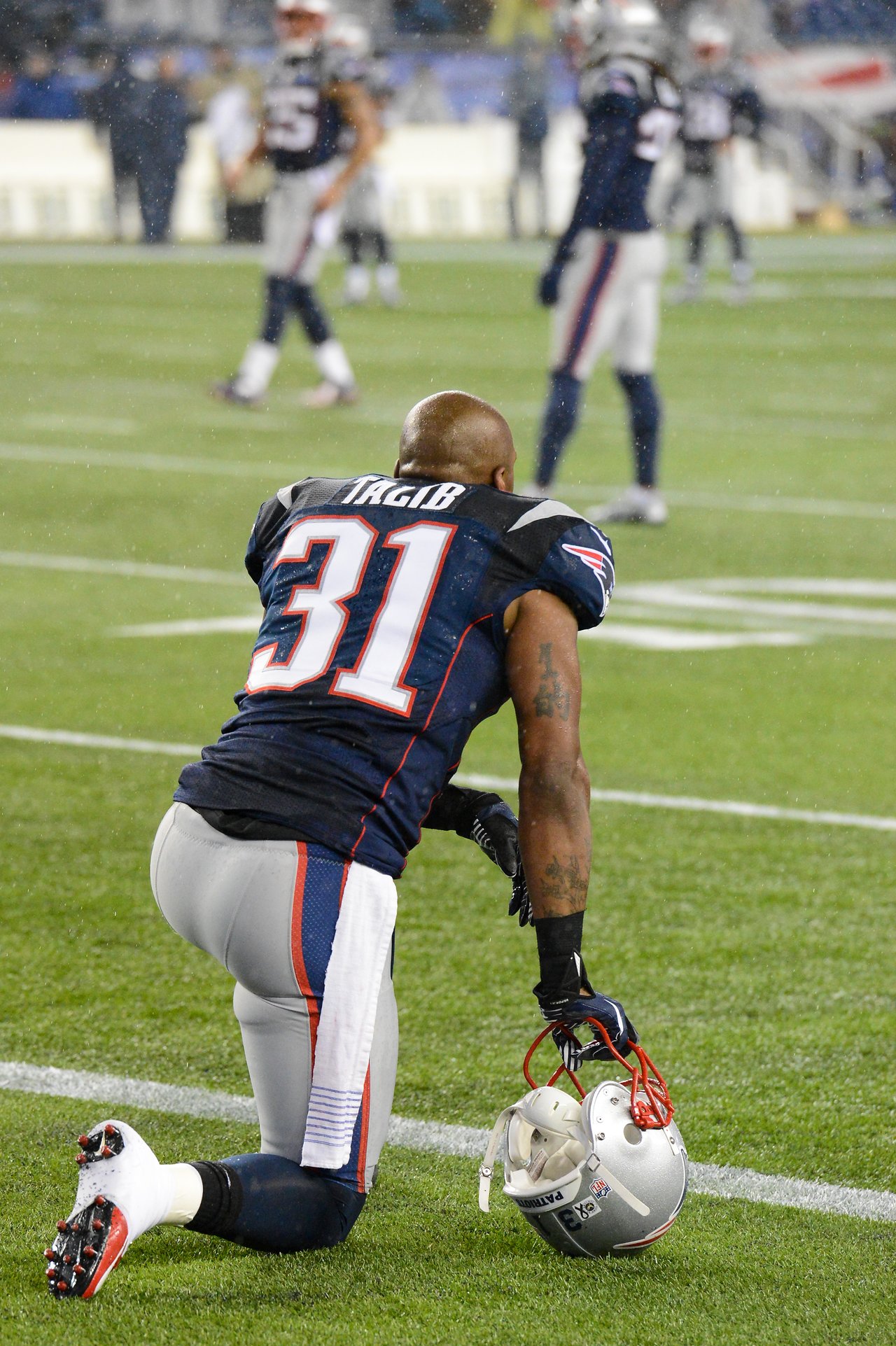 A Patriots player kneels on the field, holding his helmet, while teammates stand in the background.