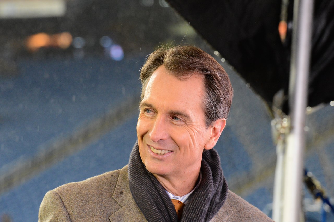 A man in a coat and scarf smiles while standing in a stadium during a Patriots game.