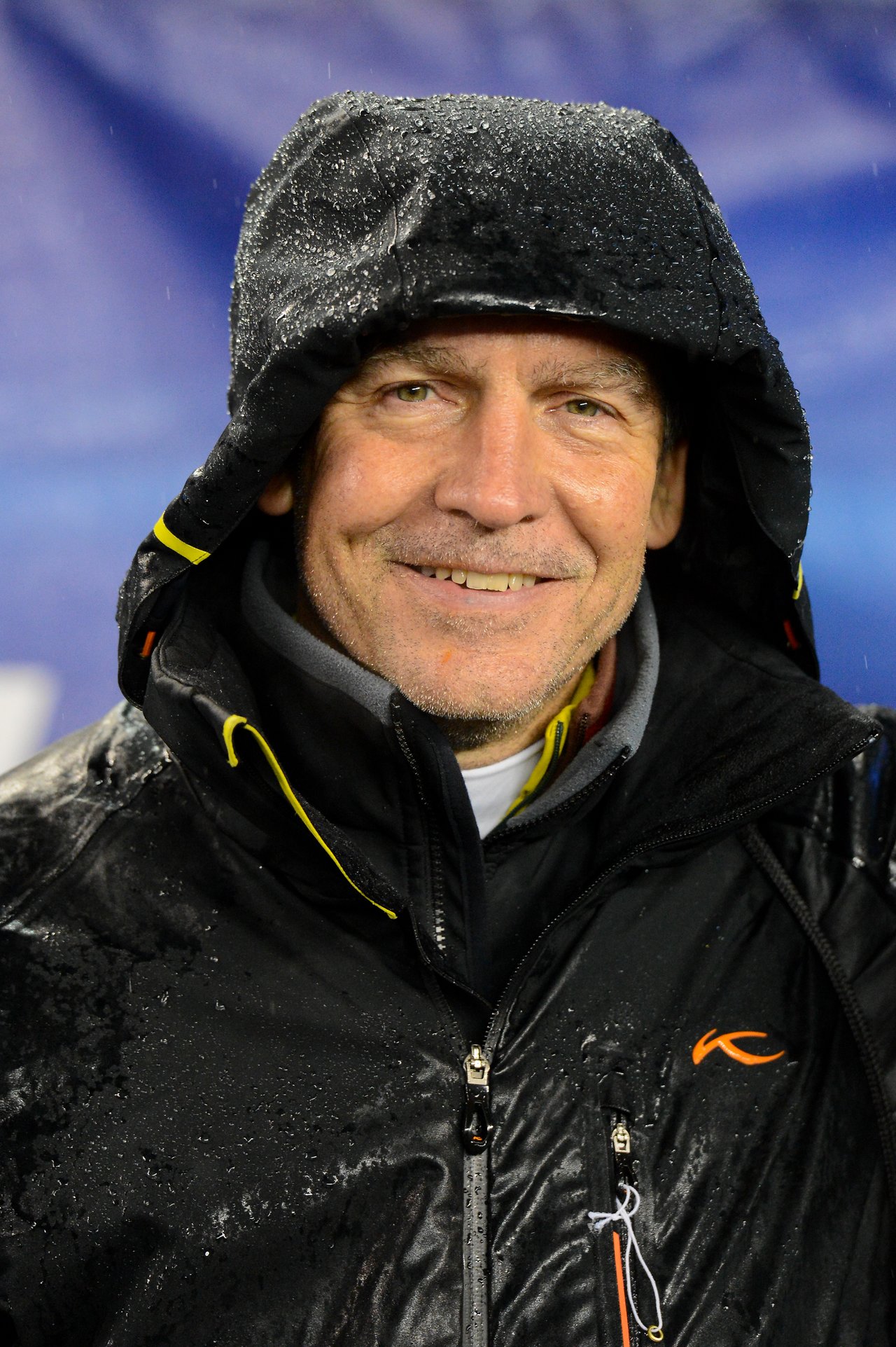 A man in a hooded rain jacket smiles while standing in the rain at a Patriots game.