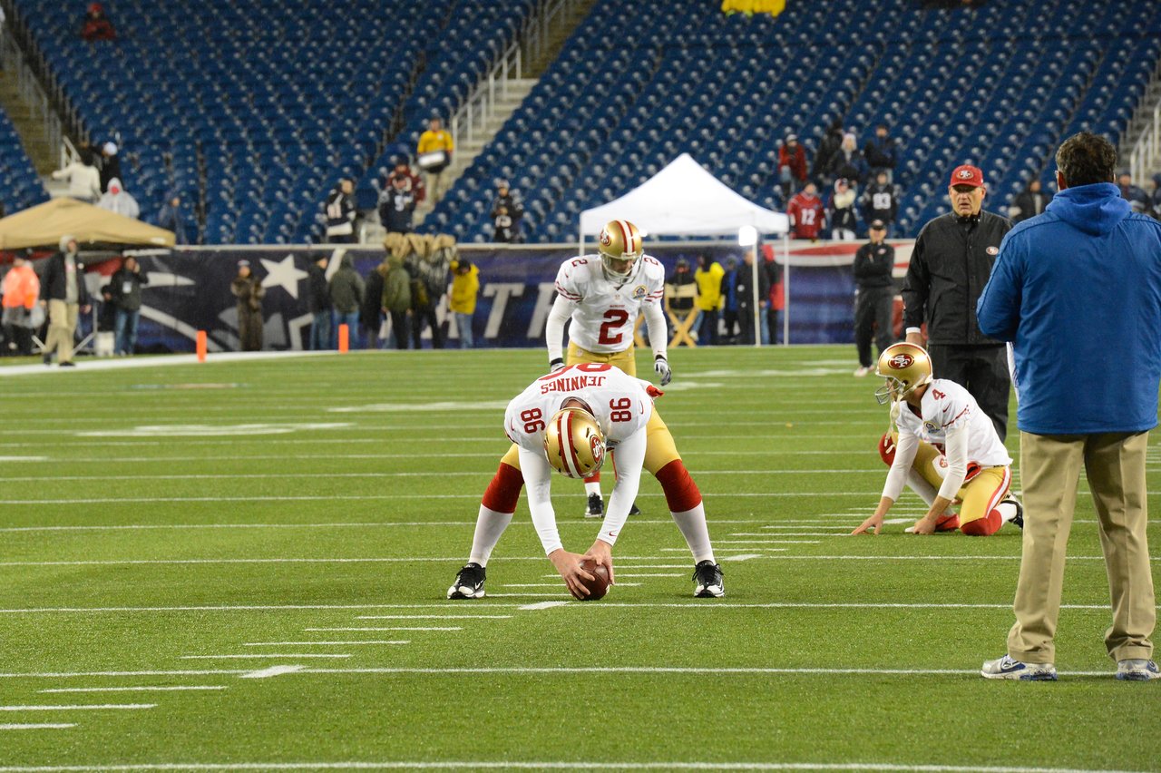 Football players in 49ers uniforms practice a field goal snap and kick before the game in a stadium.
