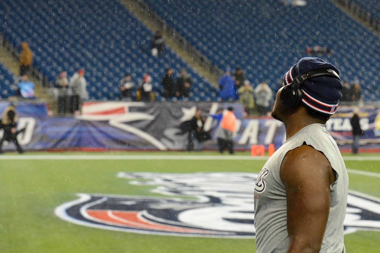 A football player wearing headphones and a beanie stands on the field before a Patriots game in the rain.
