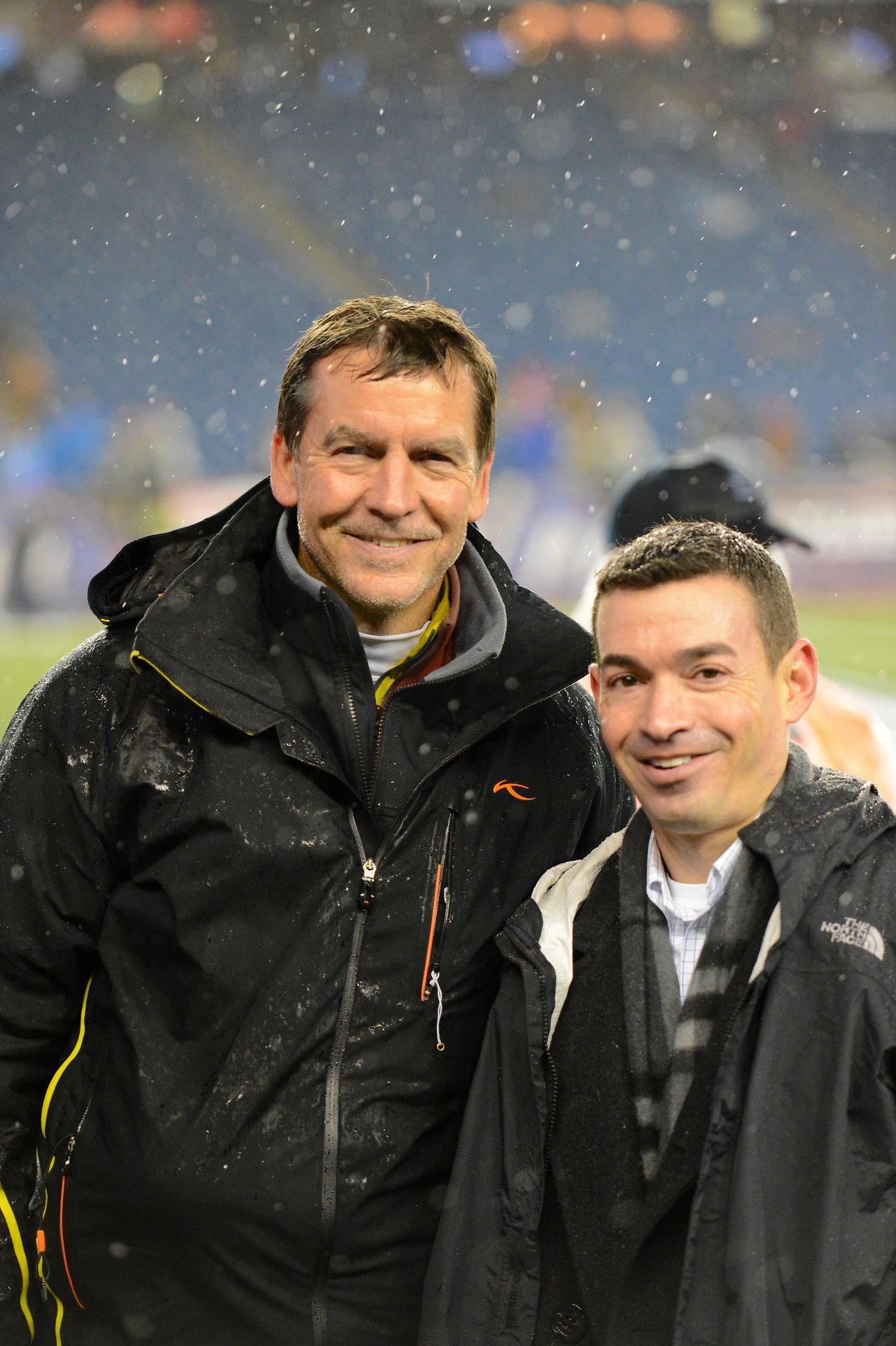 Two men in jackets smiling in a snowy stadium during a Patriots game.