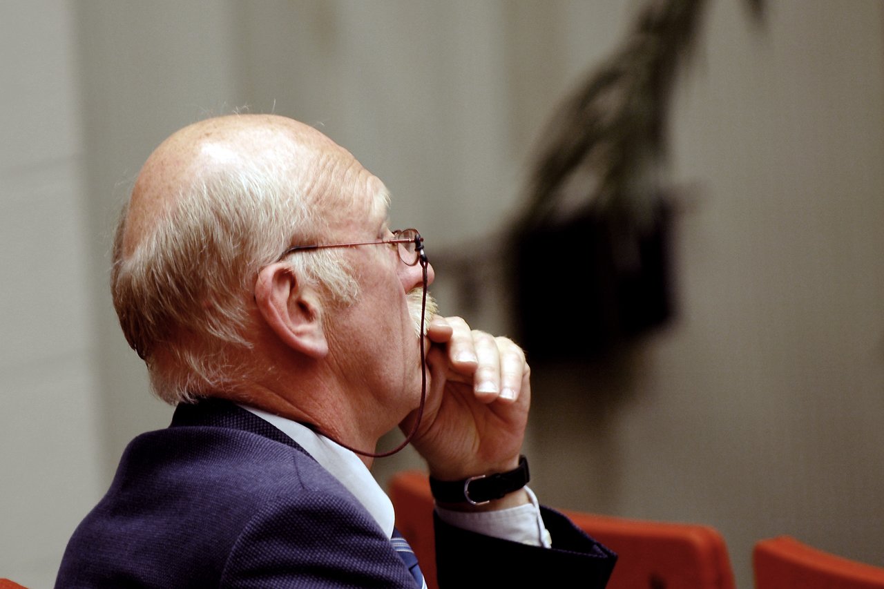 An older man in a suit holds his glasses and listens attentively during a conference or academic event.