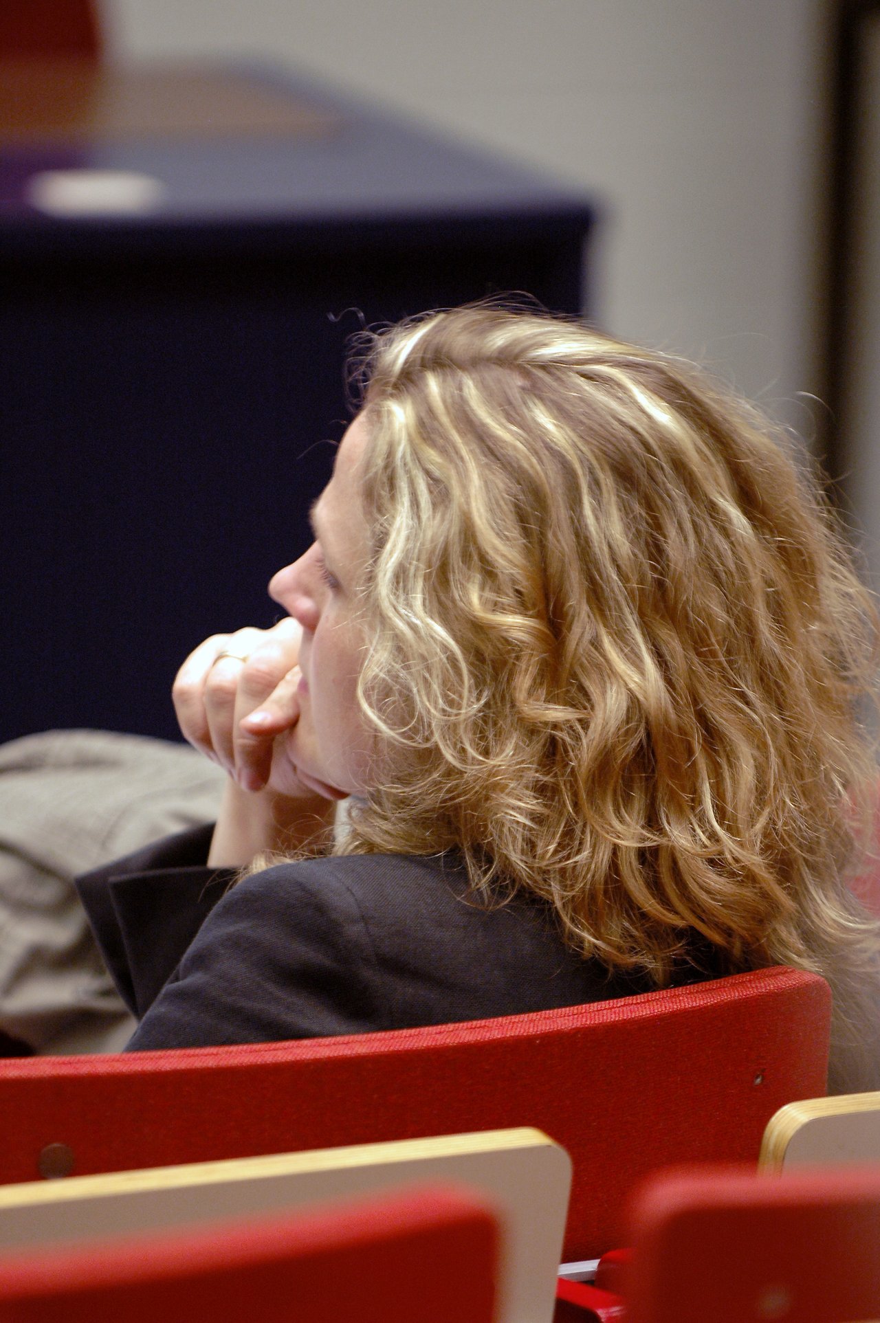 A woman with curly blonde hair sits in a red auditorium chair, resting her chin on her hand.