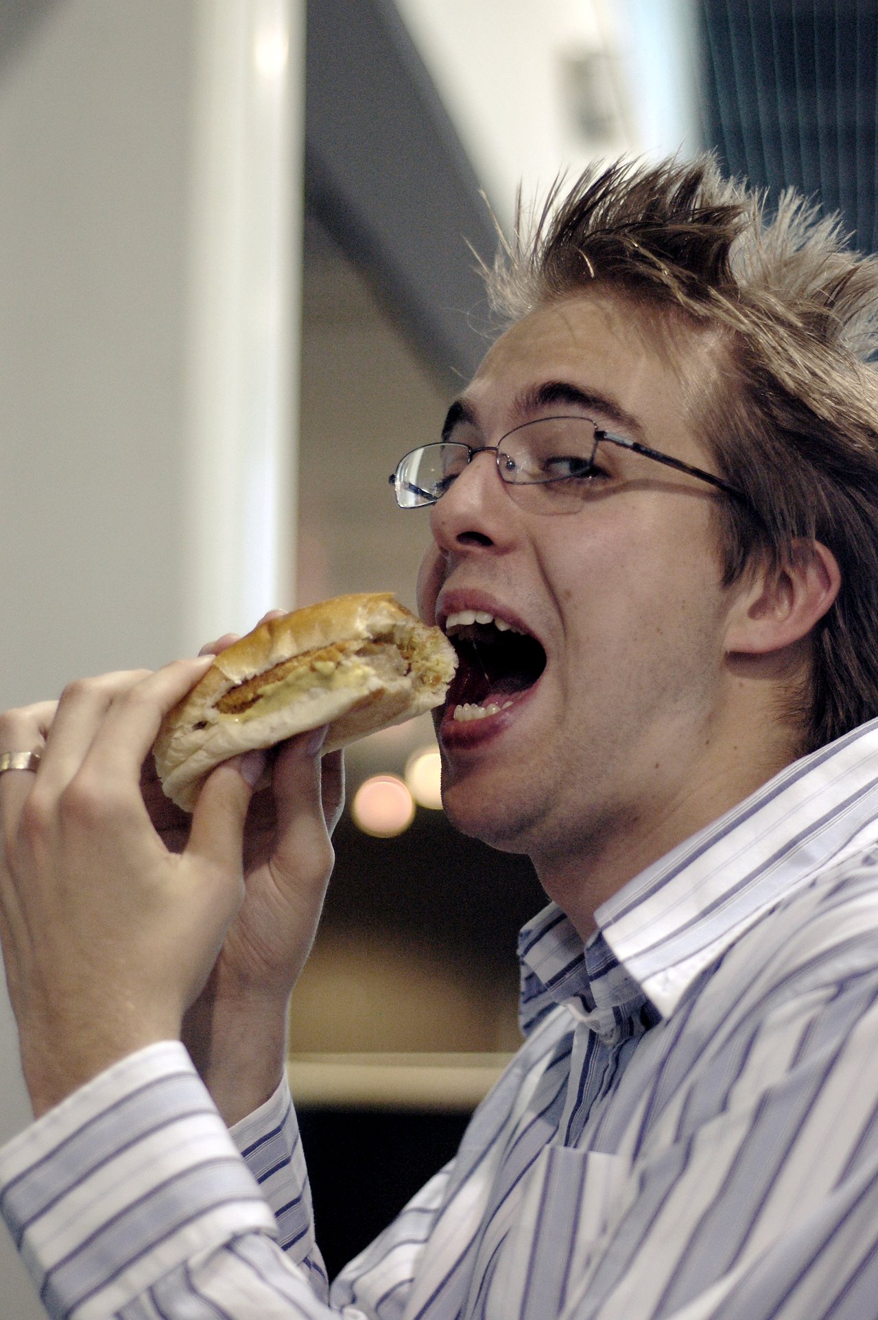 A person wearing glasses and a striped shirt takes a bite of a sandwich while looking at the camera.