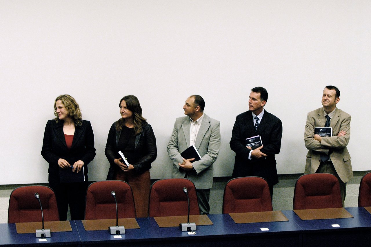 Five jury members stand in a row, holding documents, in front of a table with microphones and chairs.