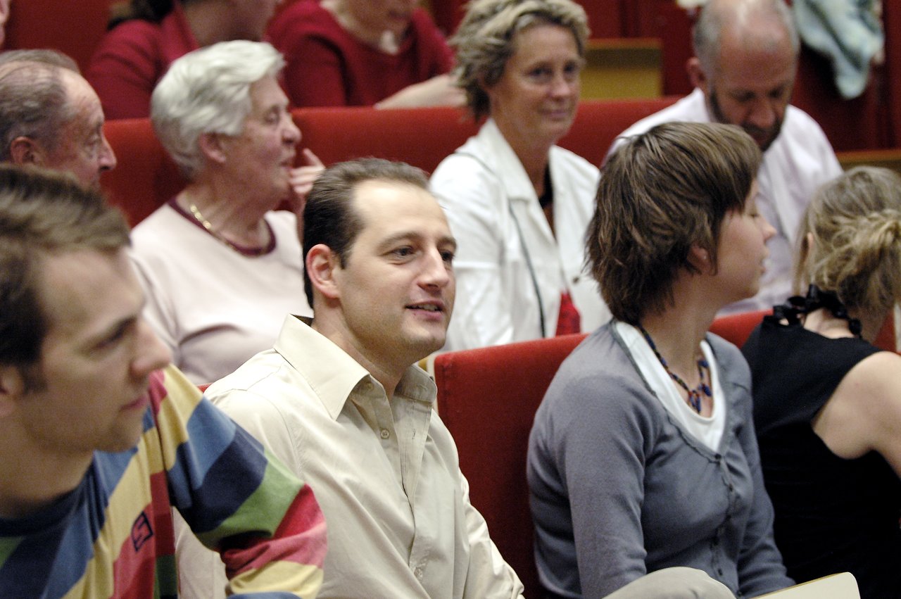 A man in a beige shirt sits in a red auditorium seat, attentively listening with others around him.