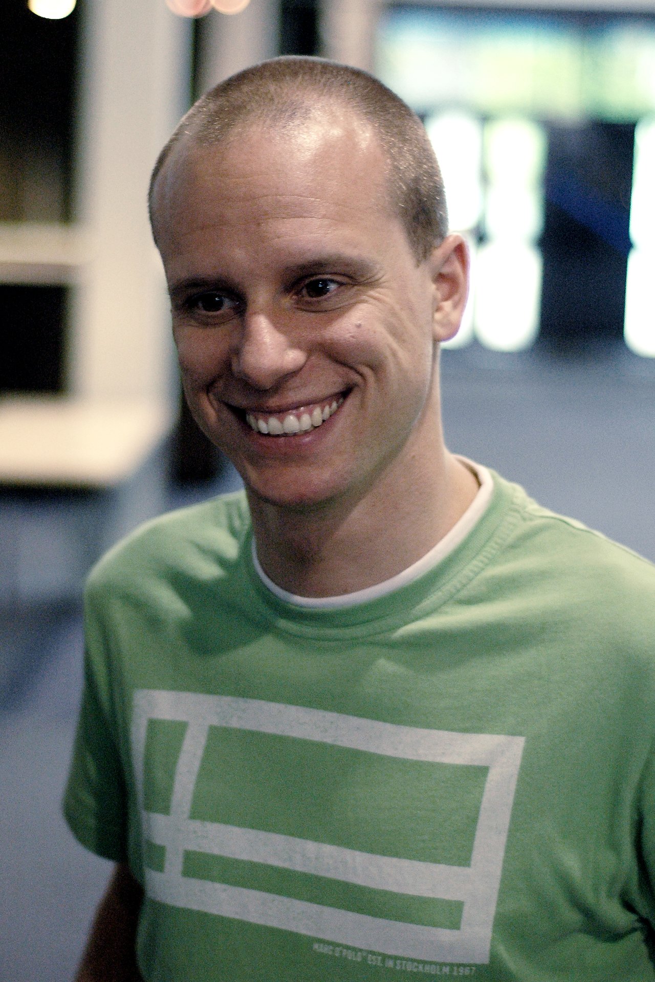 A smiling man in a green t-shirt stands indoors, looking slightly to the side.