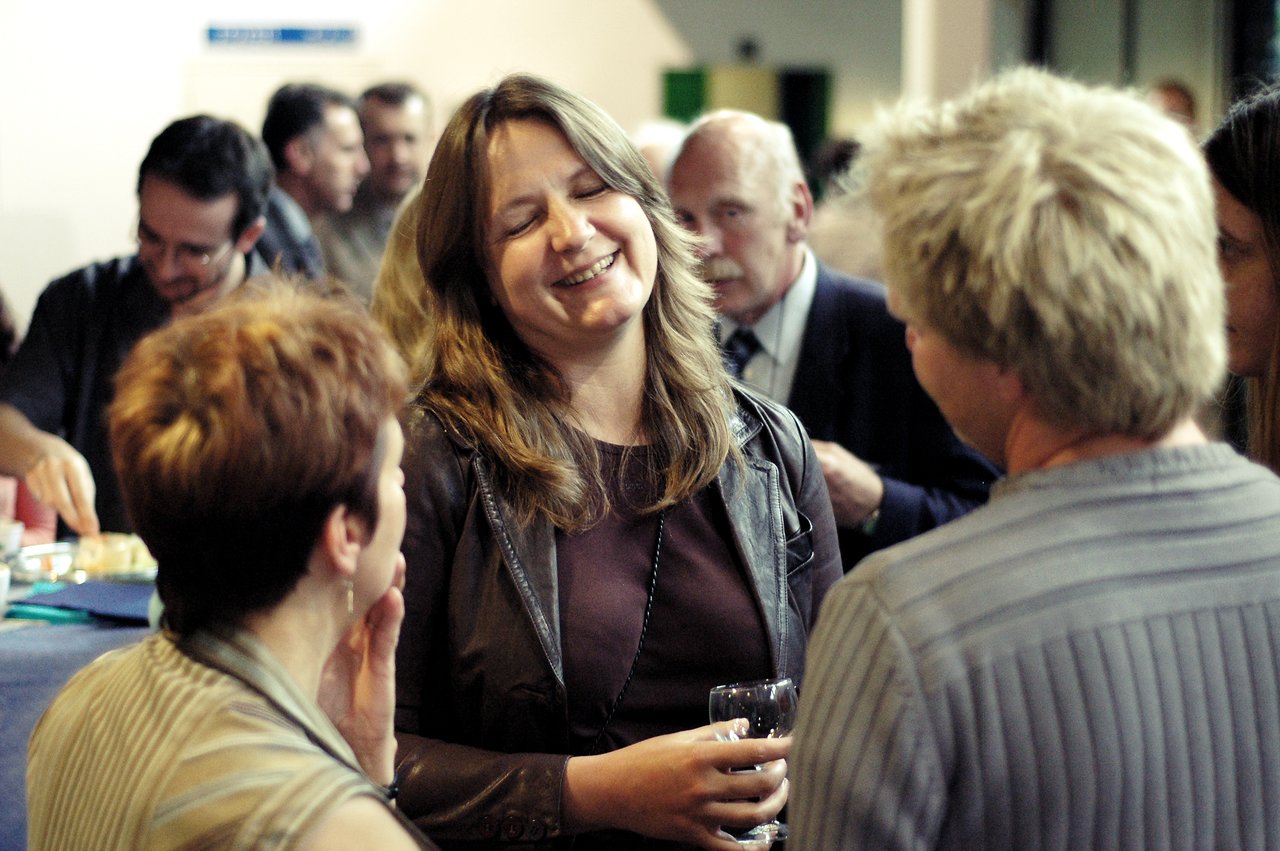A woman smiles while holding a glass and talking with two people at a reception.