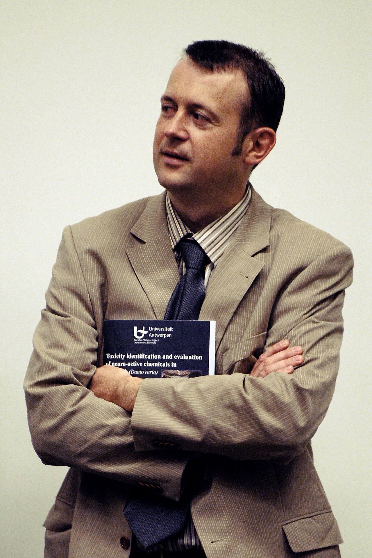 A man in a suit stands with arms crossed, holding a University of Antwerp book.