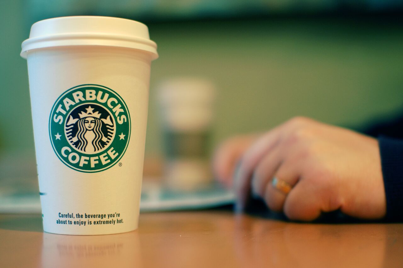 A Starbucks coffee cup on a table with a person's hand resting nearby.