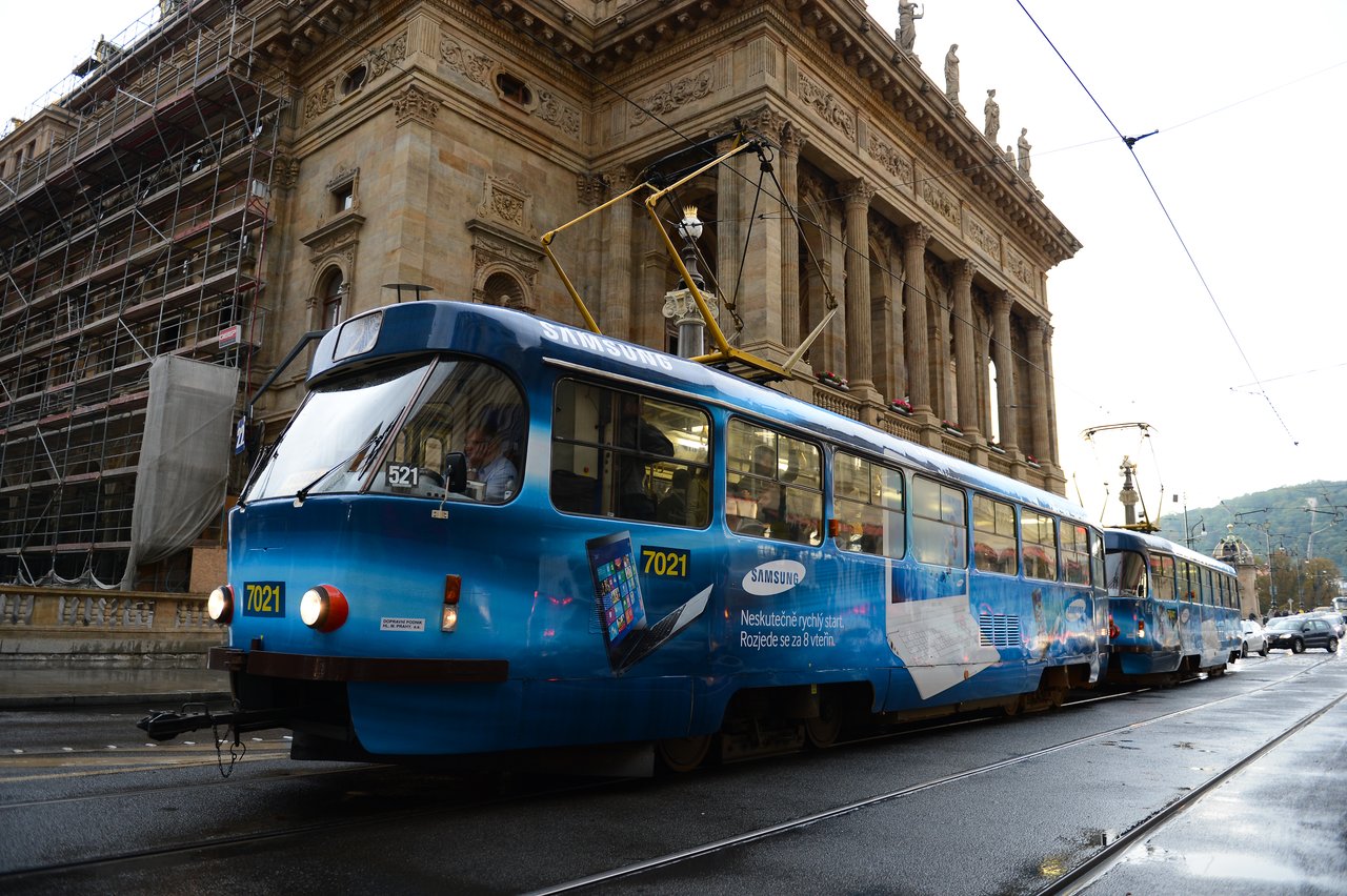 A blue tram with advertisements moves along tracks in Prague, passing a historic building under renovation.