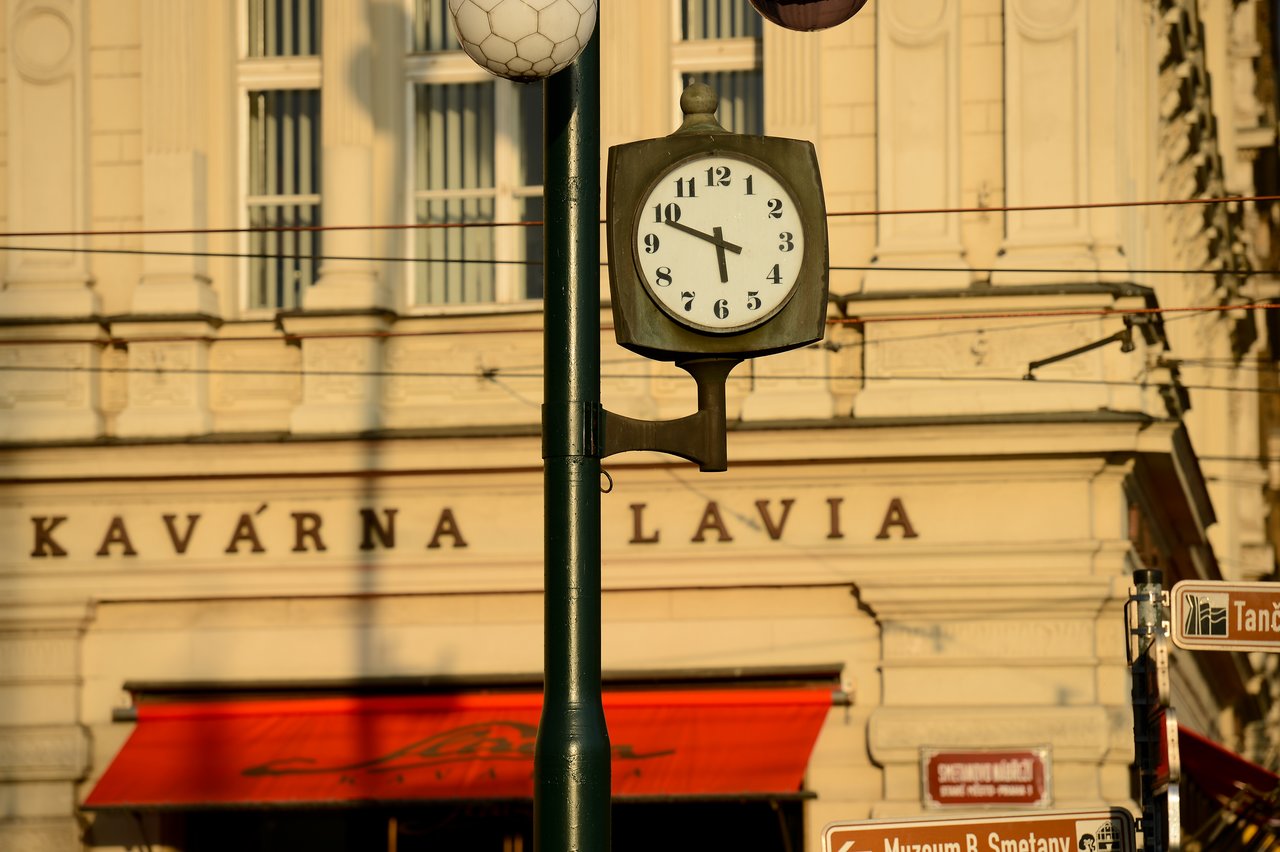 A street clock shows 4:50 in front of a café with a red awning in Prague.