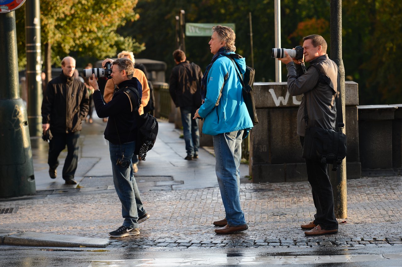 Three photographers with cameras stand on a wet sidewalk, capturing images in different directions.