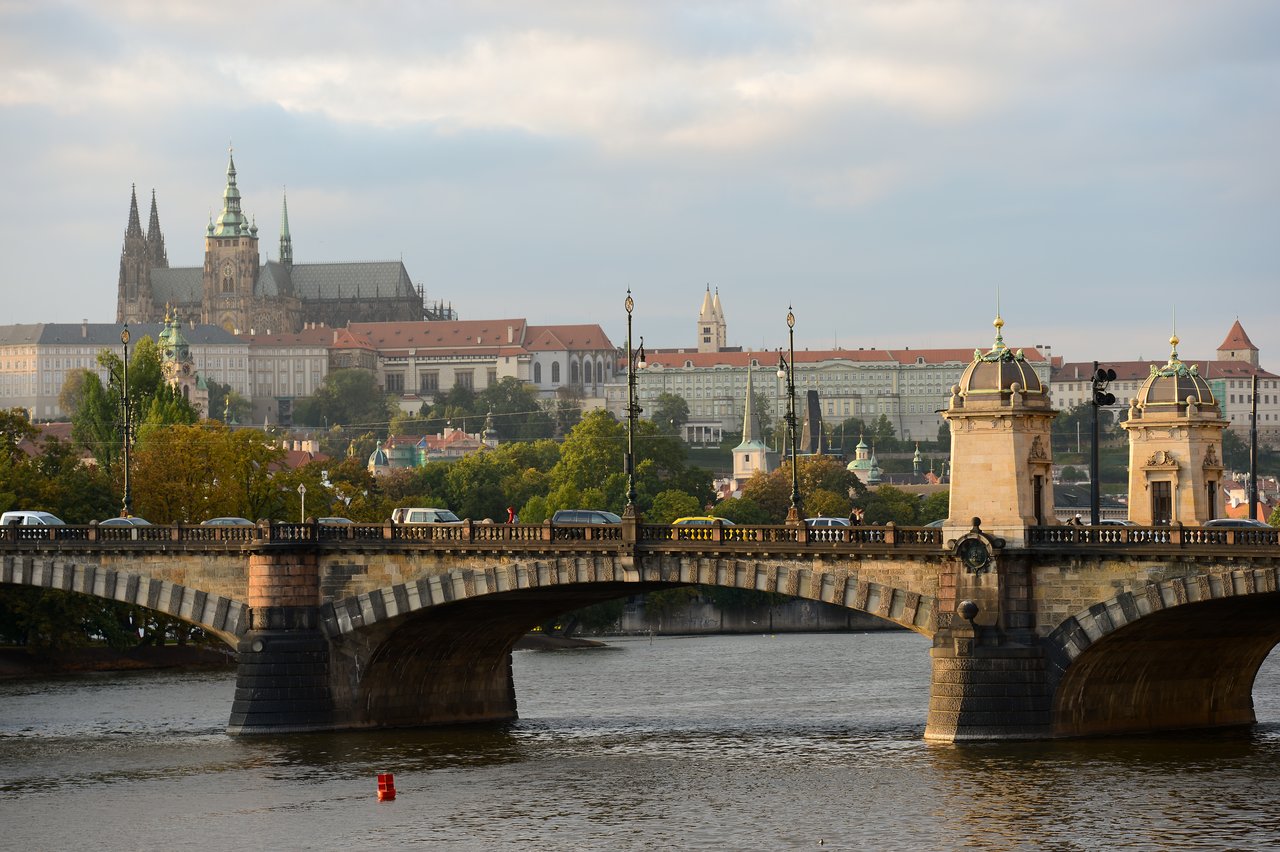 A stone bridge with cars crosses a river, with historic buildings and a large cathedral in the background.
