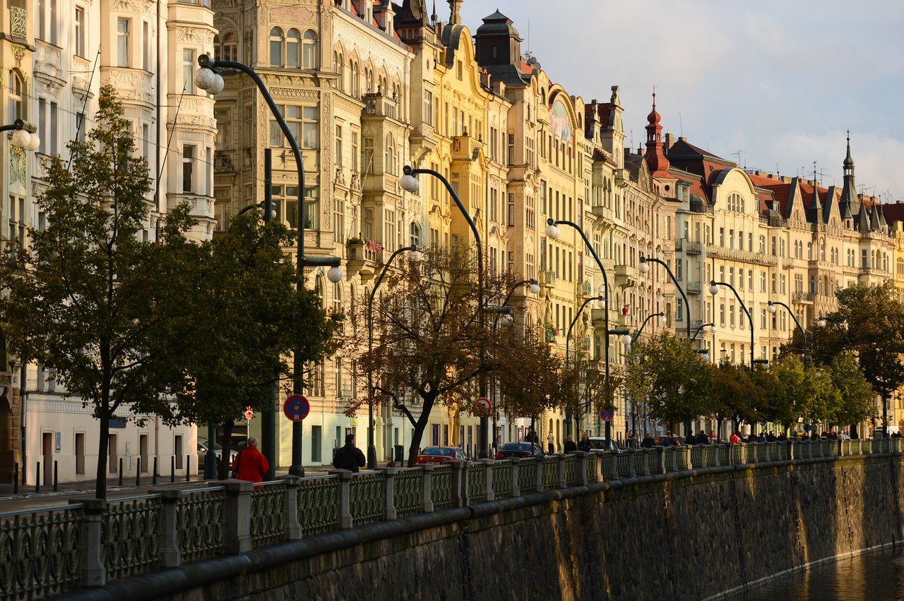 A row of historic buildings along a riverbank in Prague, with people walking on the sidewalk.