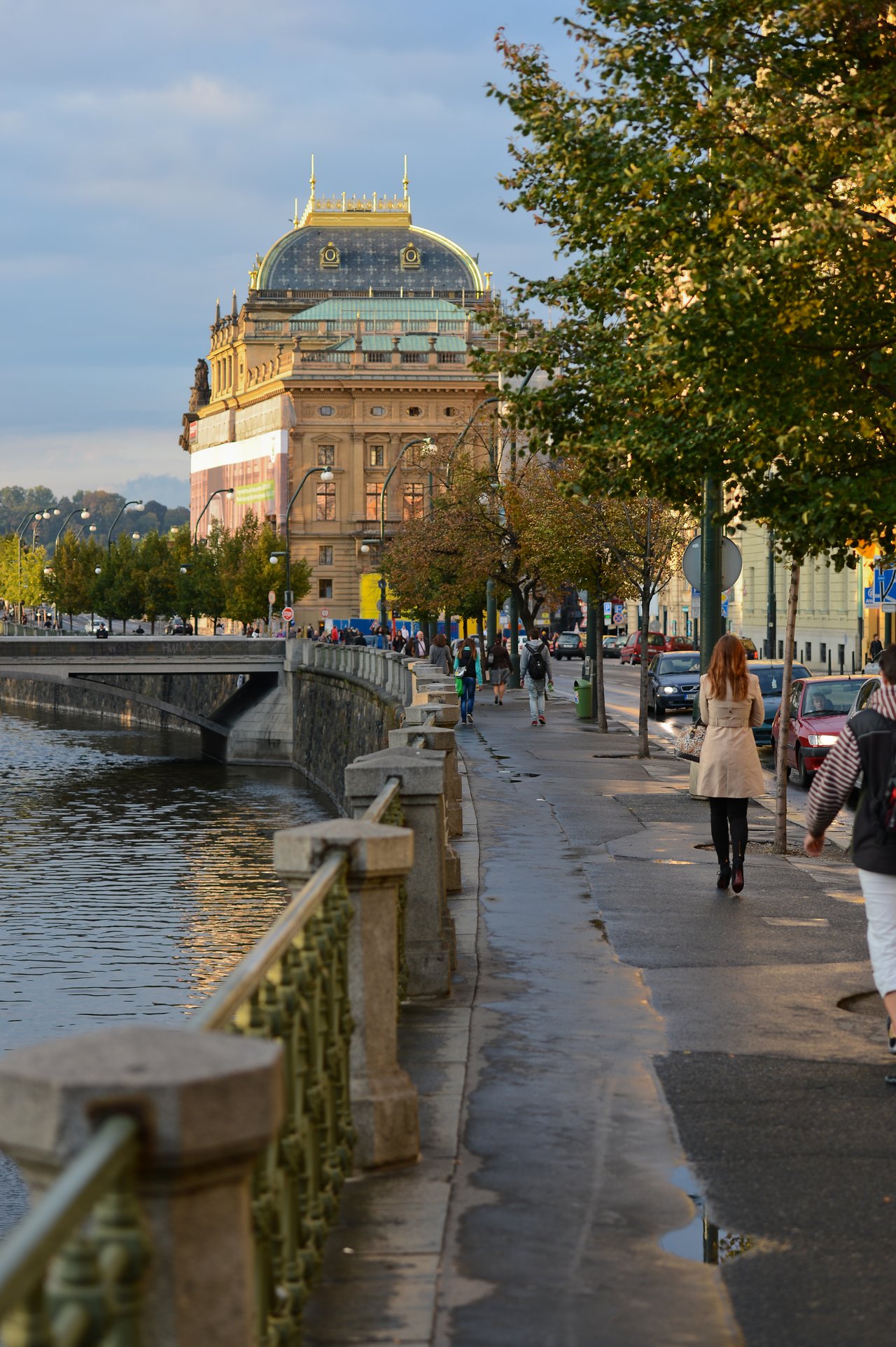 People walk along a riverside sidewalk in Prague, with a historic building in the background.