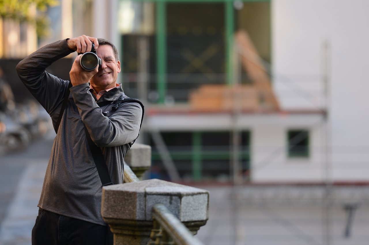 A man in a gray jacket holds a camera and takes a photo while standing near a railing.