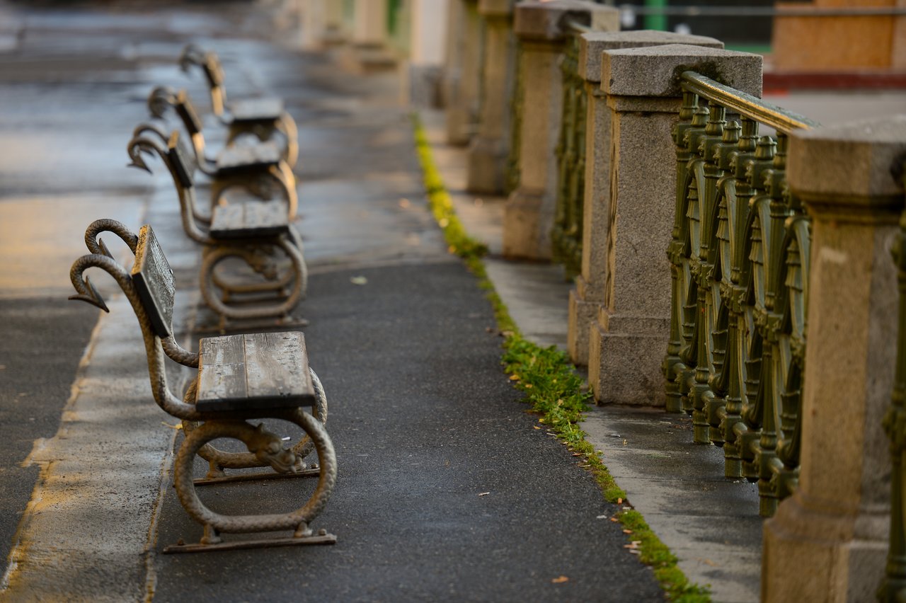 A row of empty wooden benches with metal frames lines a paved walkway beside an ornate railing in Prague.
