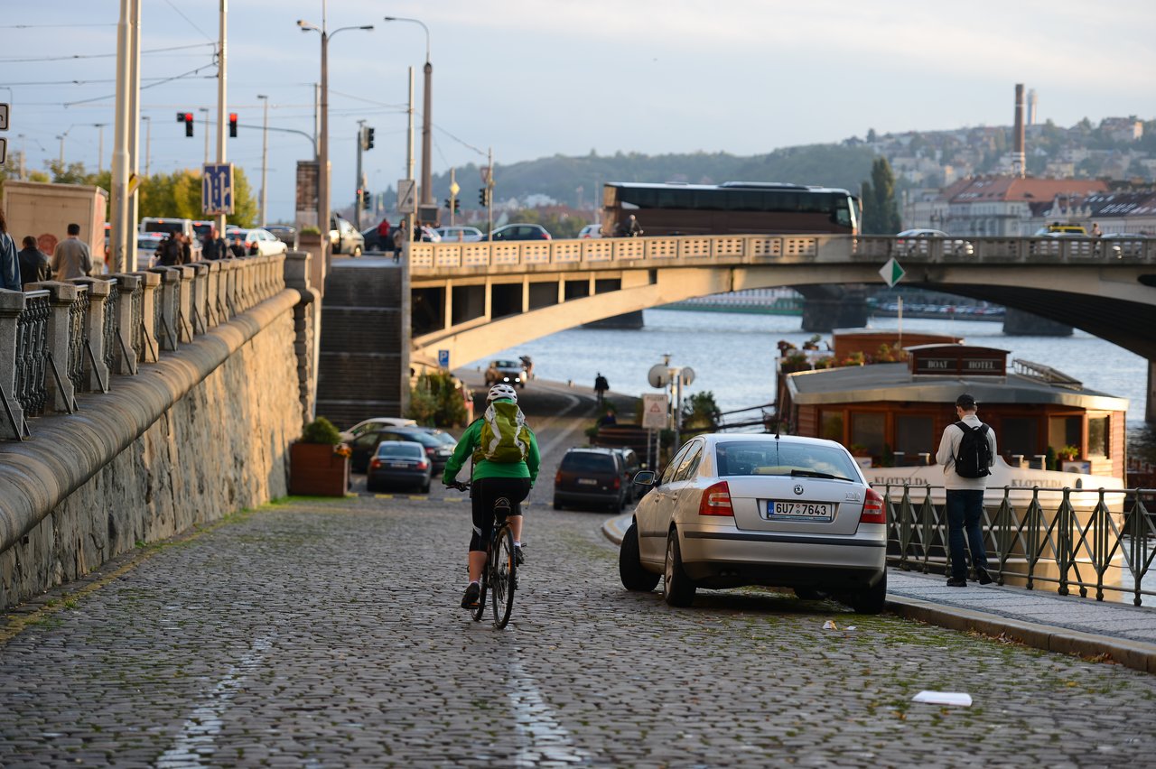 A cyclist rides on a cobblestone path near a parked car, with a bridge and river in the background.