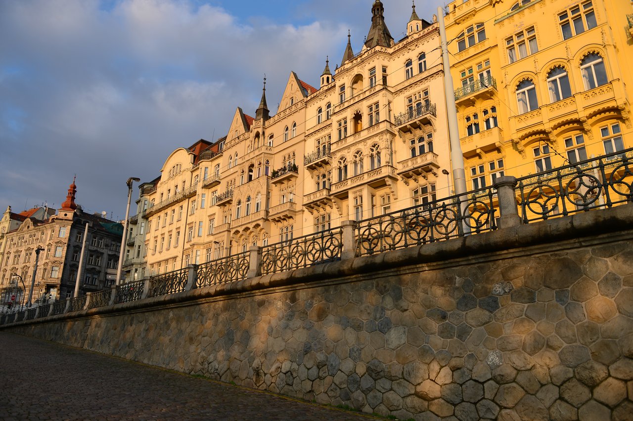 Row of historic buildings with ornate facades along a stone walkway in Prague, illuminated by warm sunlight.