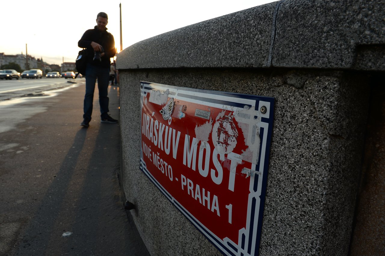 A worn street sign on a stone wall marks a location in Prague, with a person standing nearby.
