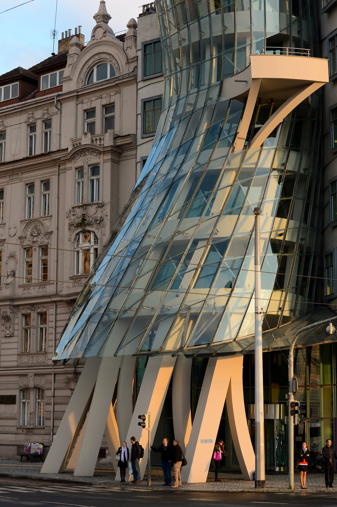 A modern glass building with a curved design, known as the Dancing House, with people walking nearby.