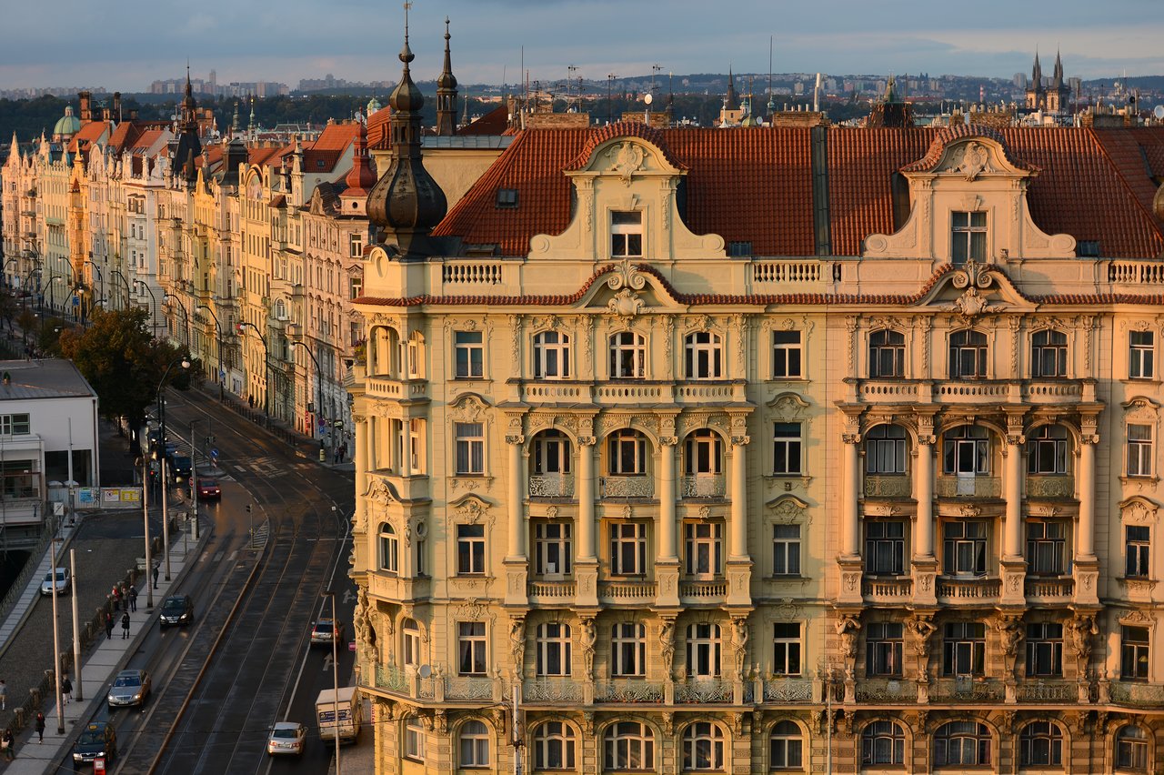Ornate historic buildings with red roofs line a curved street with tram tracks in Prague during golden hour.