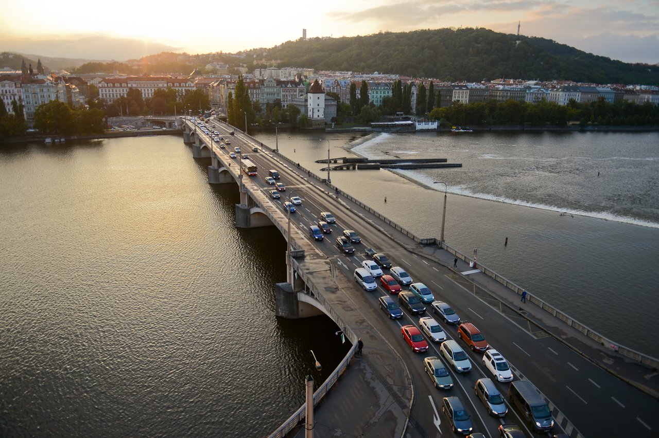 Cars in heavy traffic on a bridge over a river, with buildings and hills in the background at sunset.
