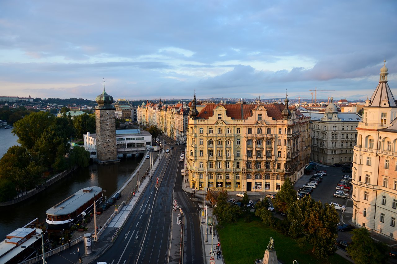 A cityscape of Prague with historic buildings, a river, a tower, and a busy street with cars and pedestrians.