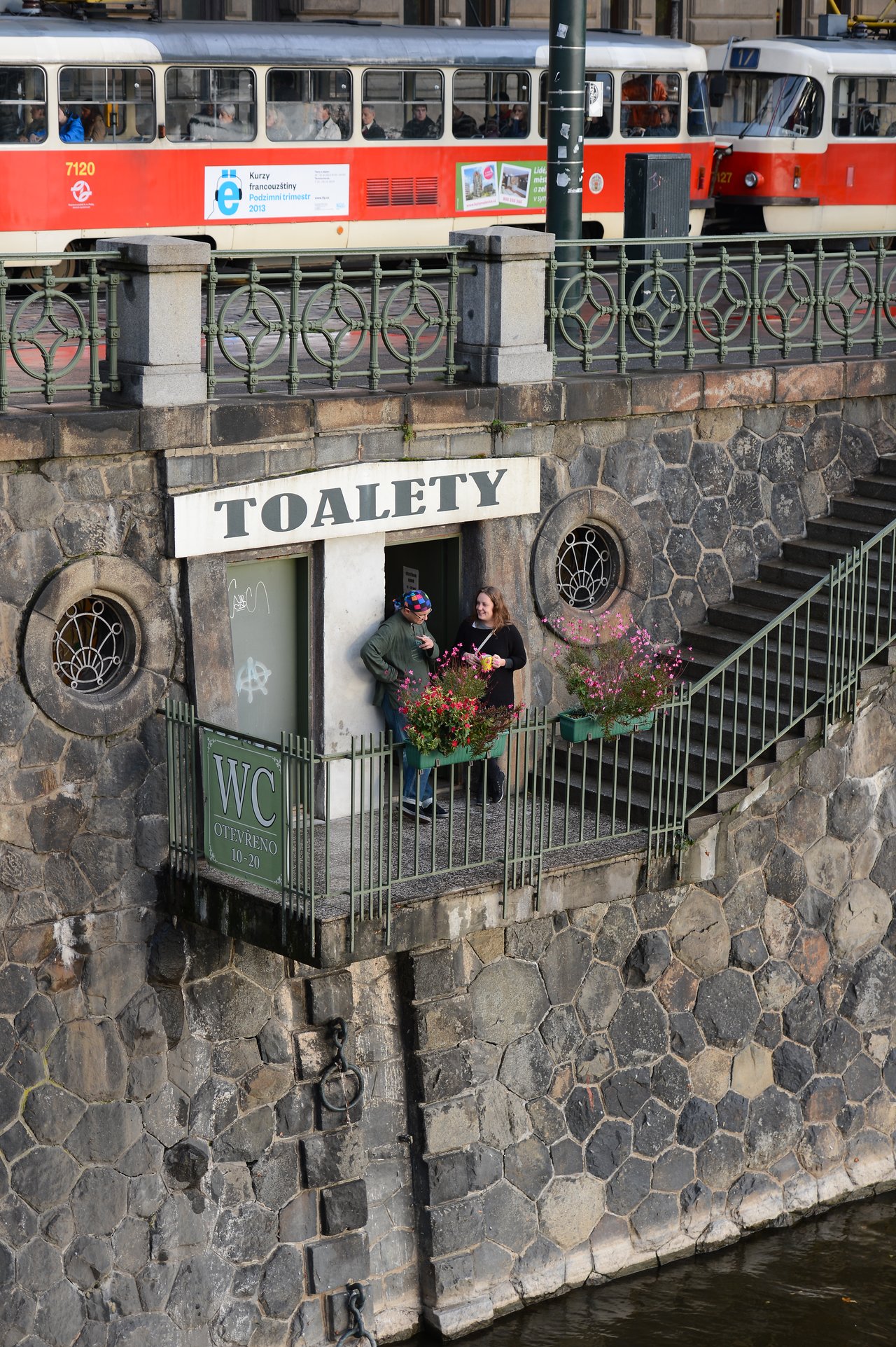 Two people stand outside a public restroom entrance on a stone wall, with trams passing on the street above.
