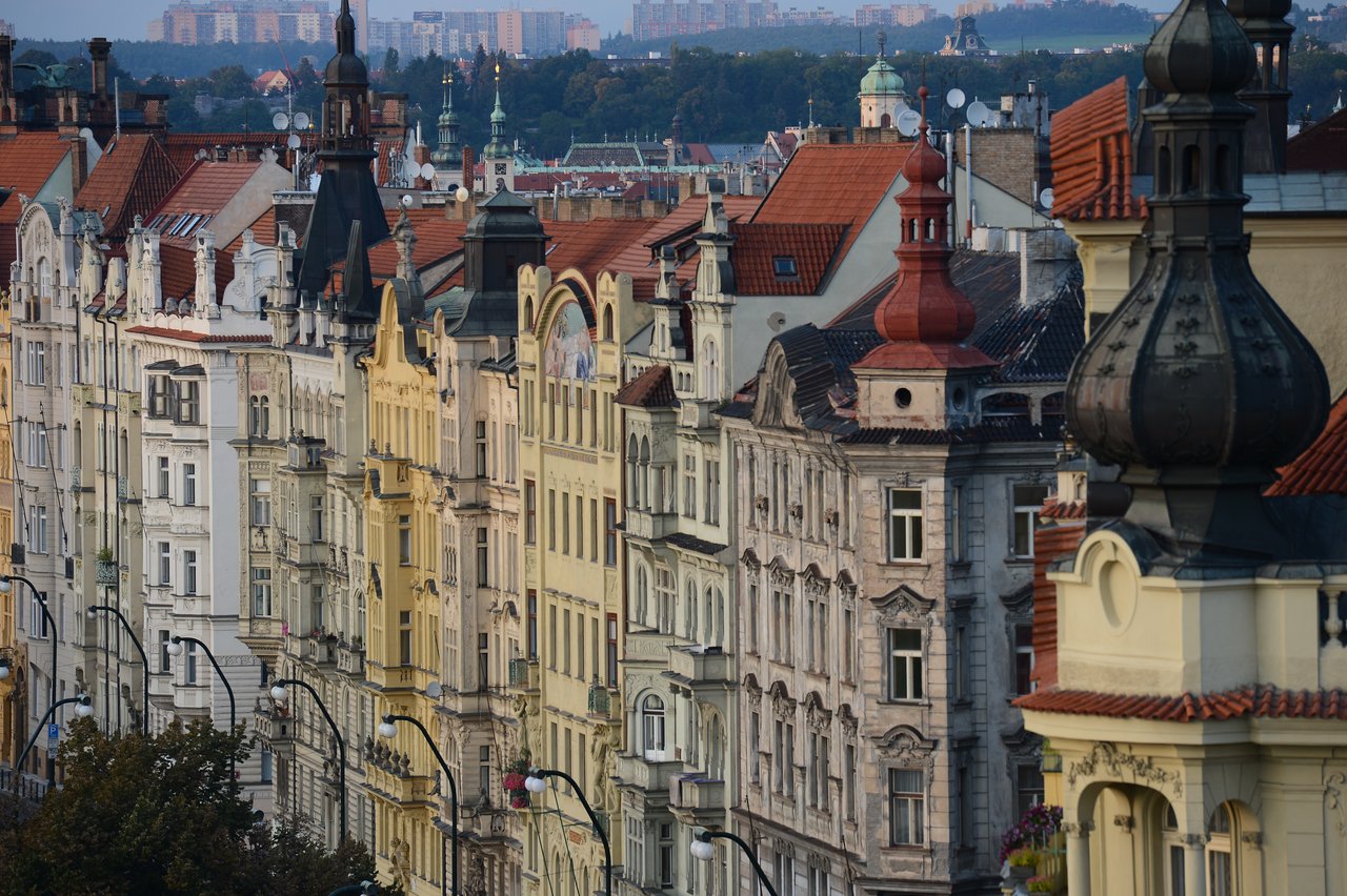 A row of historic buildings with ornate facades and red rooftops in Prague, with street lamps in view.