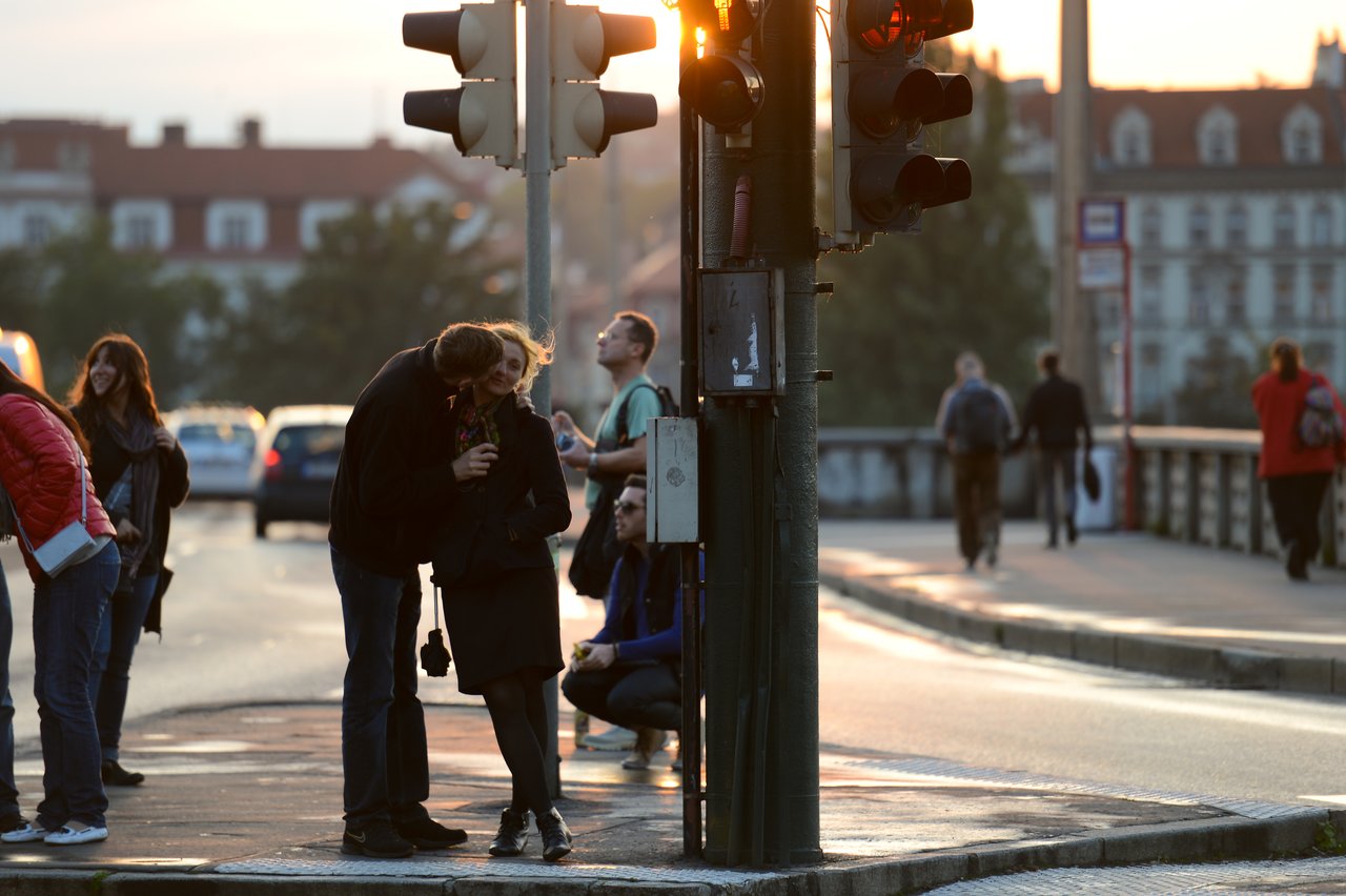 A couple stands near a traffic light, with one person leaning in close to the other on a busy street.