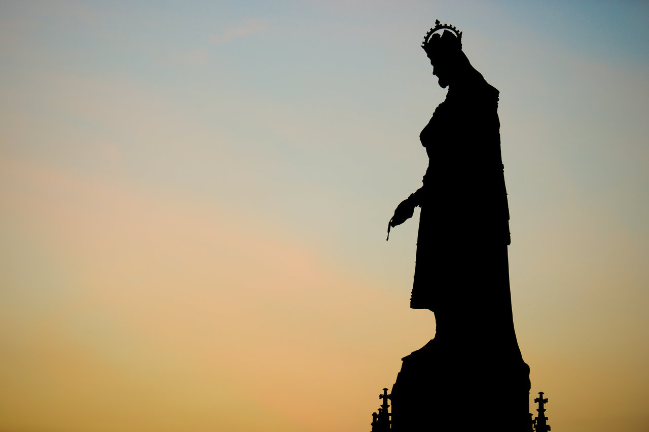 Silhouette of a crowned statue holding a small object, set against a soft, colorful sky in Prague.