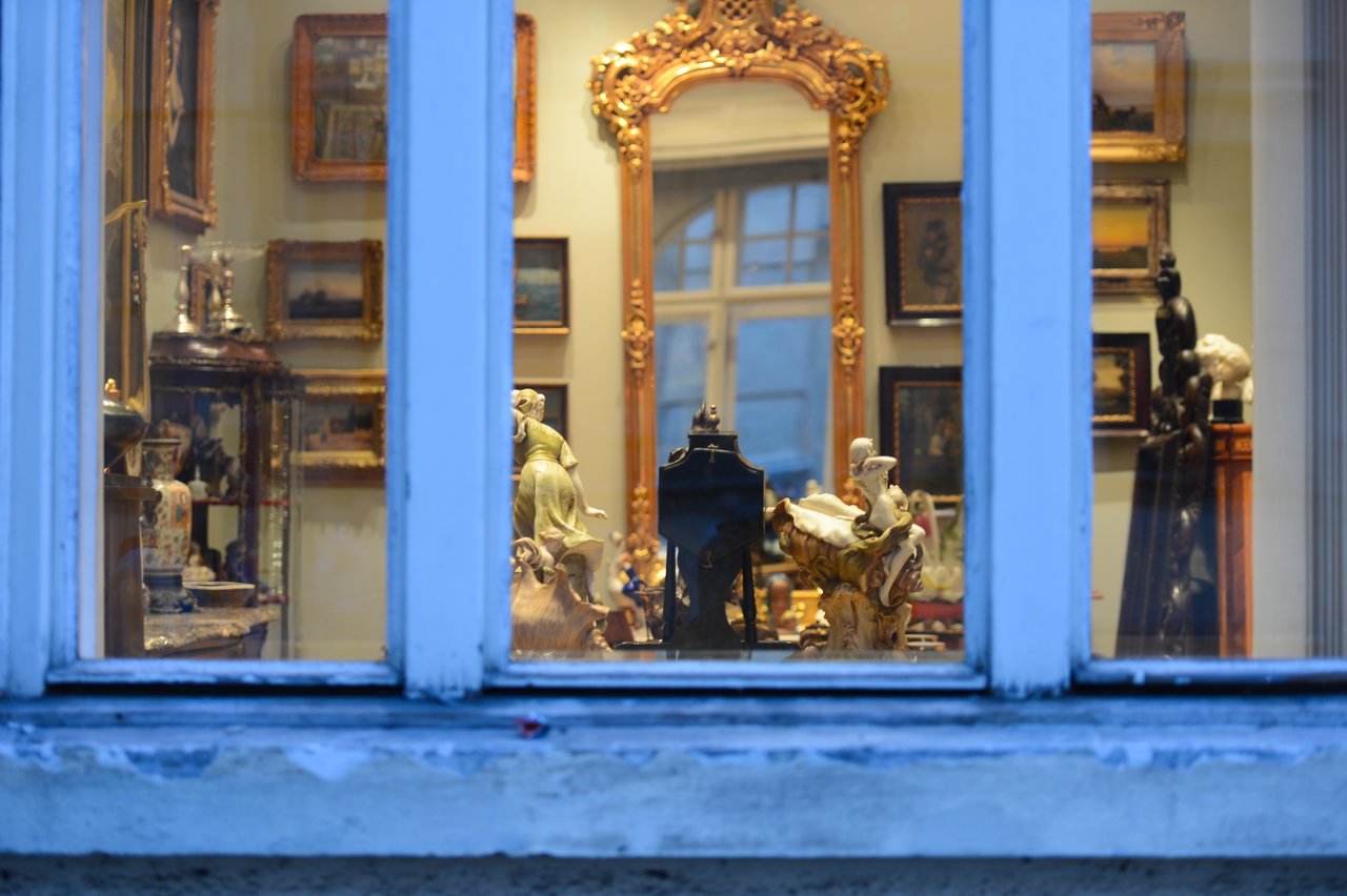 A window view of an antique shop displaying framed paintings, sculptures, and decorative items inside a well-lit room.