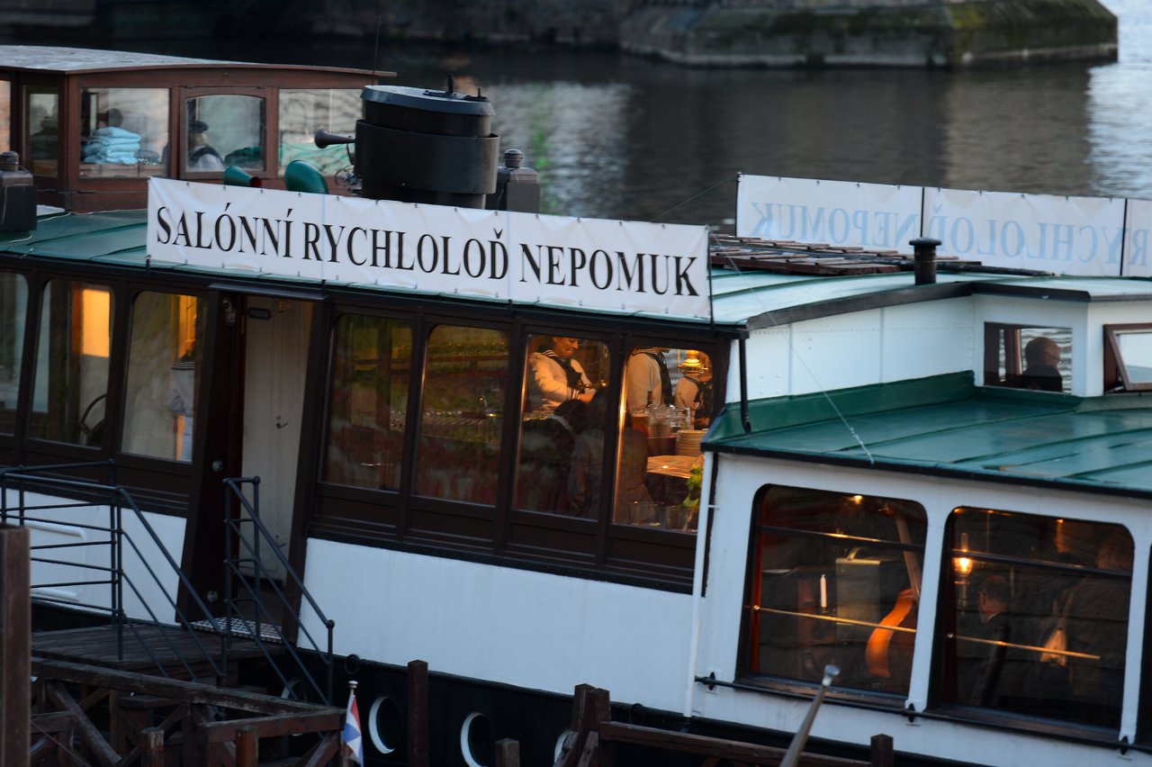 A riverboat with large windows docked by the water, with people dining inside under warm lighting.
