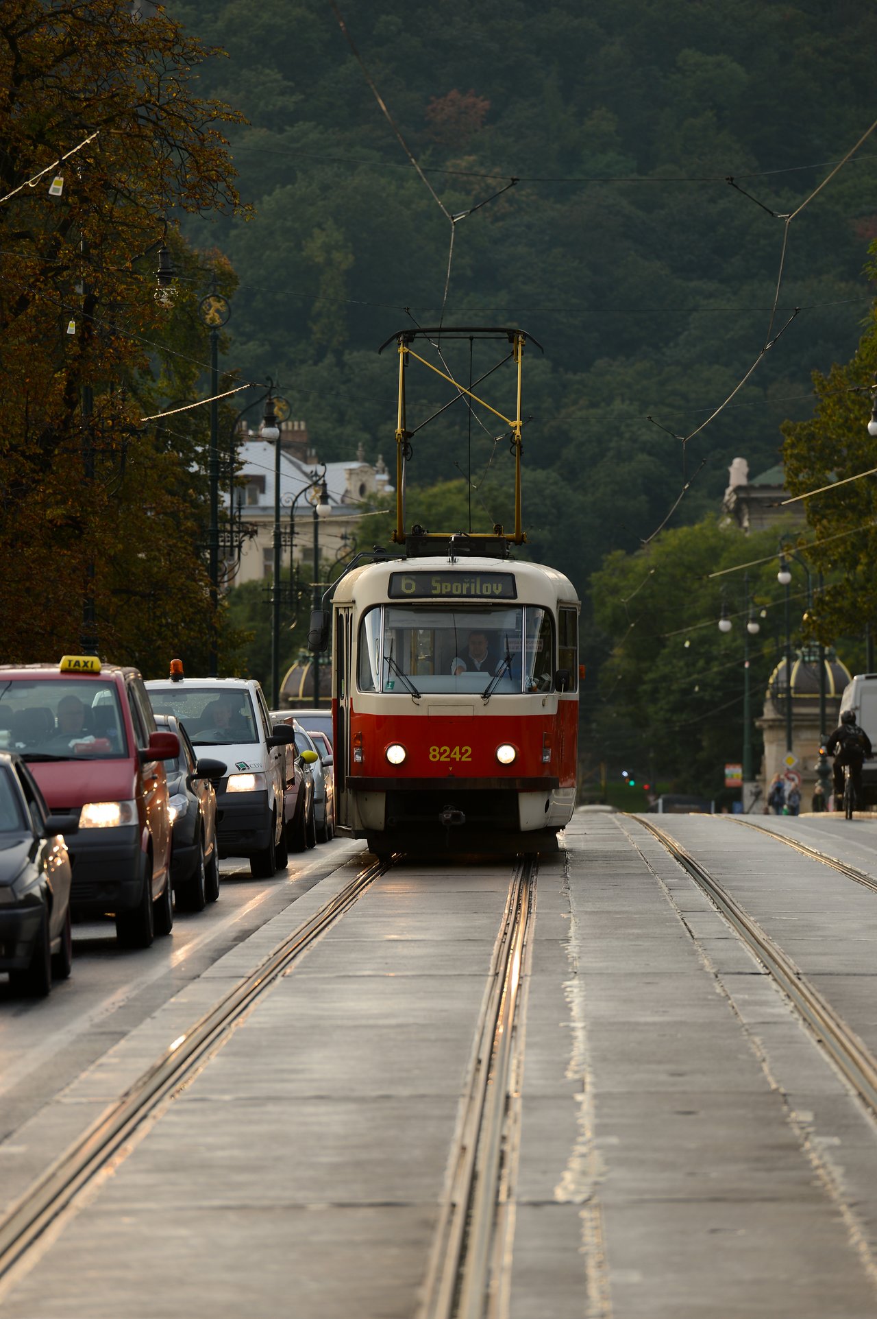 A red and white tram moves along tracks in Prague, surrounded by cars on a busy street.
