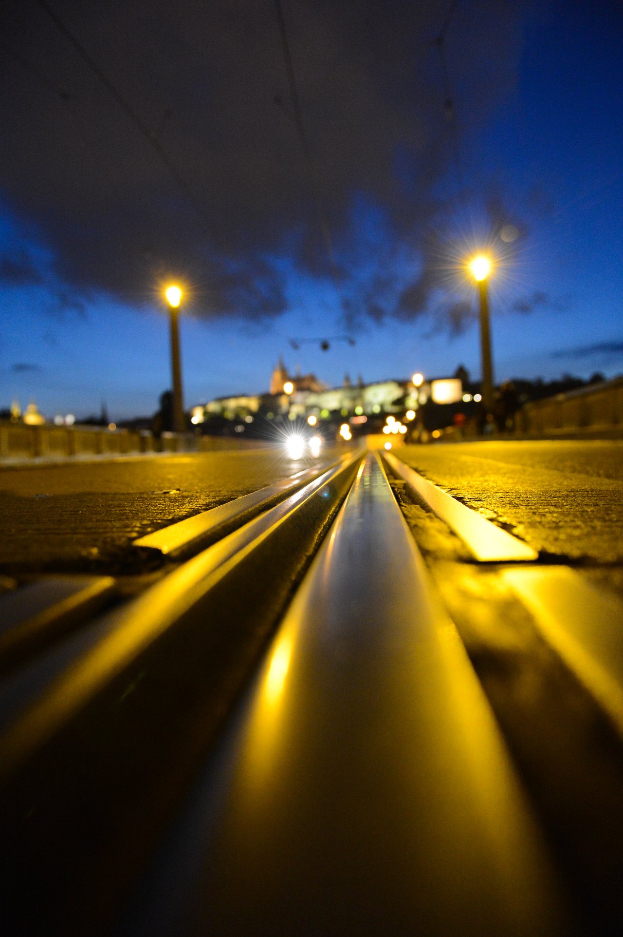 Tram tracks on a bridge at night, leading toward a lit historic building in the distance.