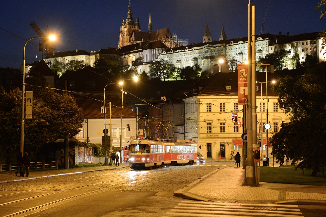 A red tram moves through a well-lit street at night with Prague Castle illuminated in the background.