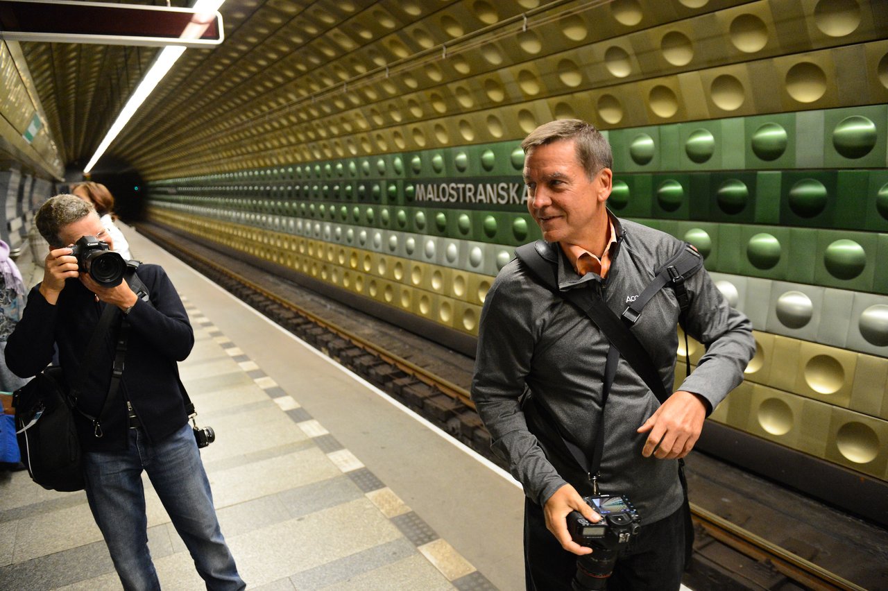 A man with a camera smiles while another person takes a photo at a Prague subway station.