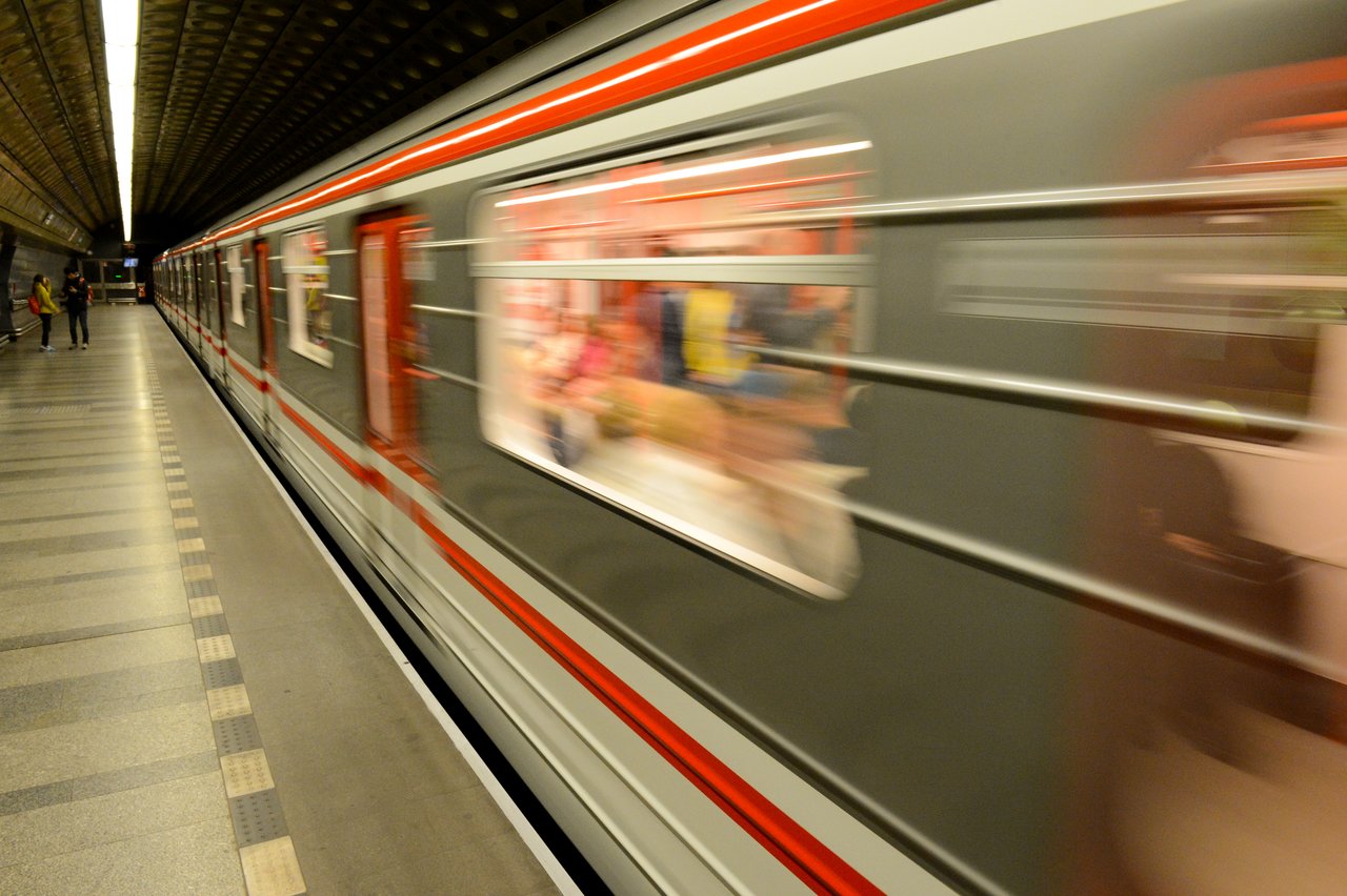 A subway train in motion passes through an underground station, with a few people waiting on the platform.