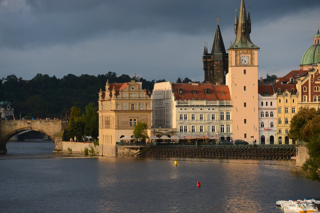 A historic clock tower and colorful buildings line the riverbank in Prague, with a stone bridge in the background.