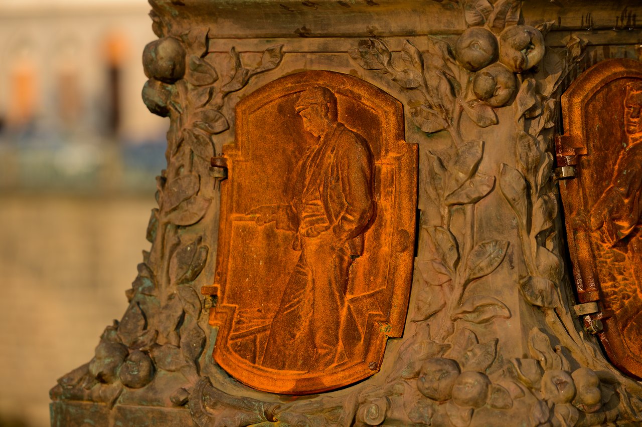 A close-up of a metal relief on a monument, showing a figure in profile surrounded by decorative leaves and fruit.