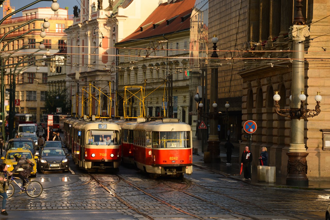 Two red and white trams move through a busy street in Prague, surrounded by cars, cyclists, and pedestrians.