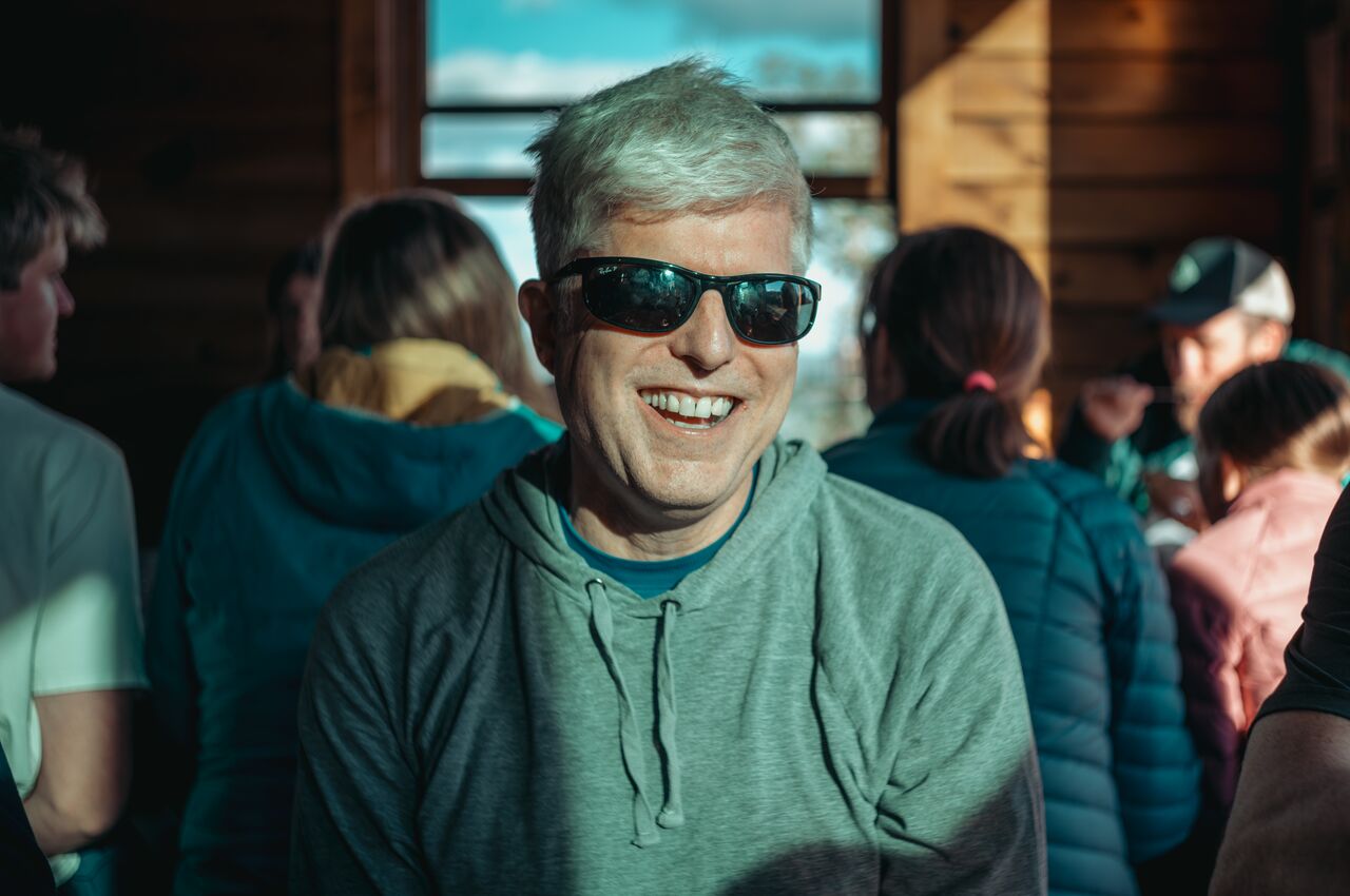 Smiling man with gray hair wearing sunglasses sitting between people inside a wooden building.